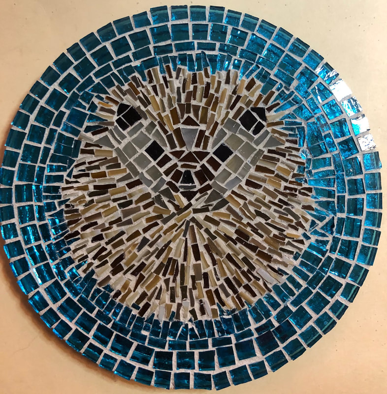 A mosaic of a cute hedgehog in a ball surrounded by blue glass after grouting.